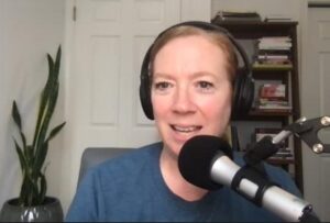 Image of a white presenting woman with a plant and bookcase in the background. The woman wears headphones, is smiling and speaking into a microphone.