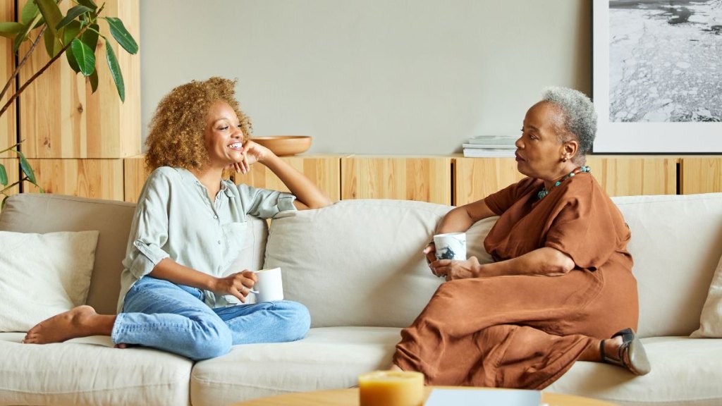 Full length of women talking with each other while sitting on sofa in living room. Mother and daughter enjoying coffee at home. They are in casuals.