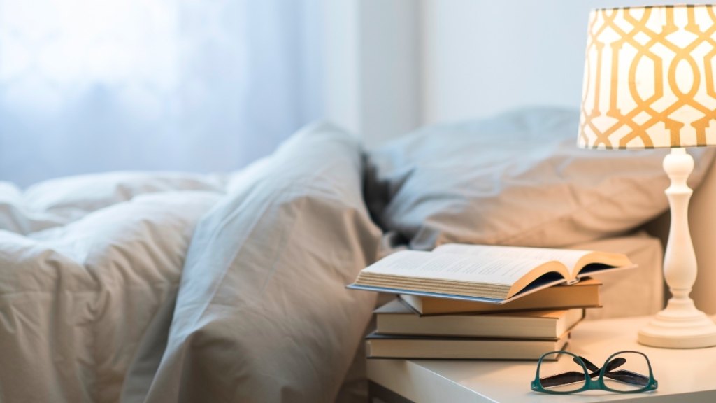 A stack of books on a nightstand with glasses an a lit lamp