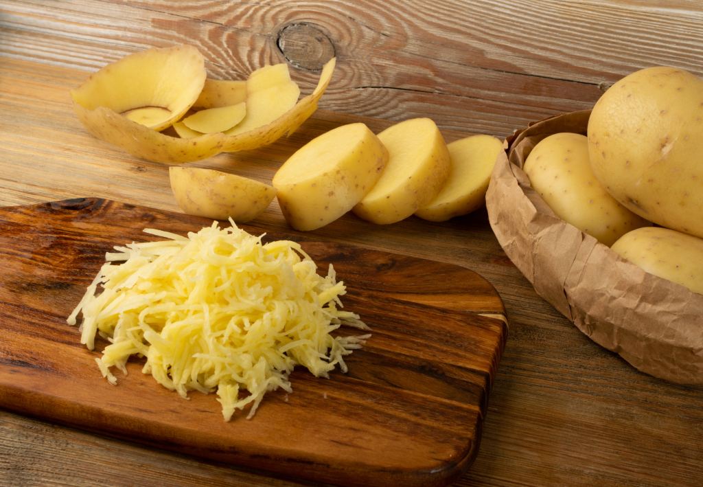 Raw grated potato on wooden cutting board background. Grate and sliced potatoes pile for swiss potato or pancakes on vintage chopping board closeup