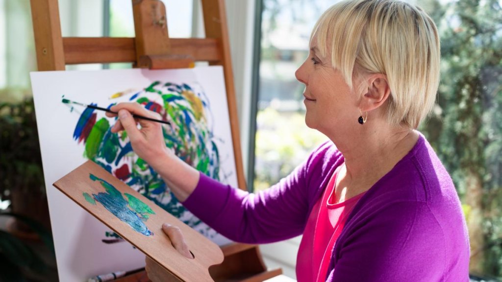 Happy retired woman painting on canvas for fun at home