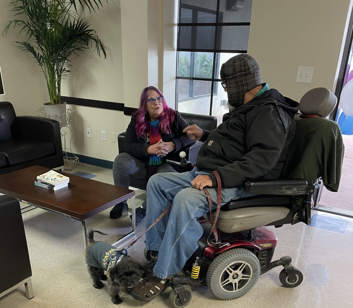 Man in wheelchair with dog talking with woman in chair. Inside, with plant and coffee table.