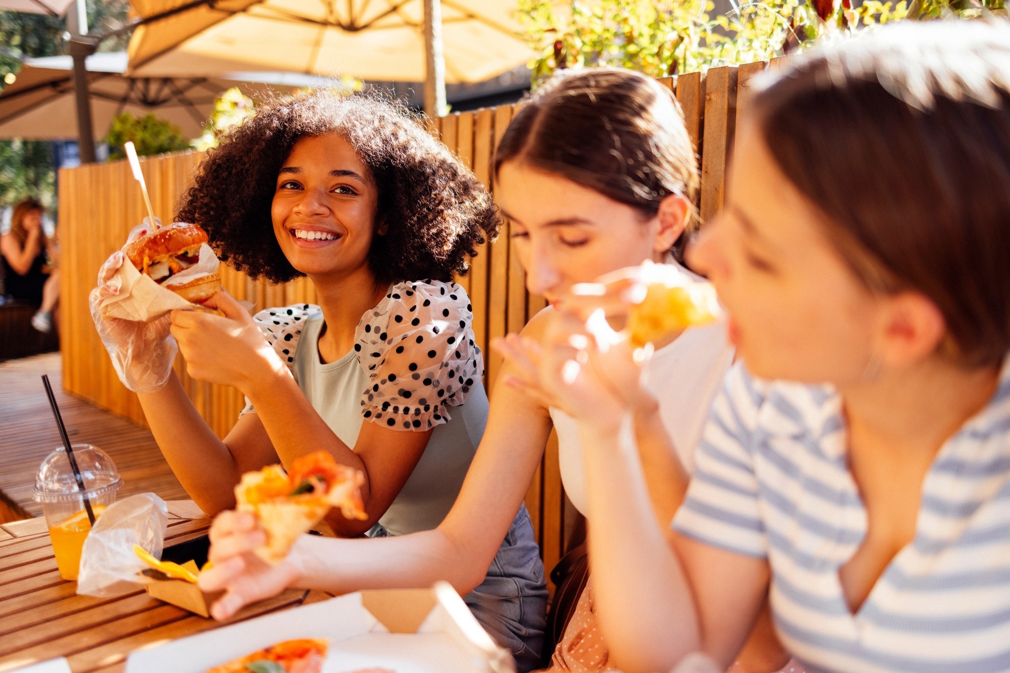 Cute smiling teenage girls are sitting in open air cafe and eating fast food.