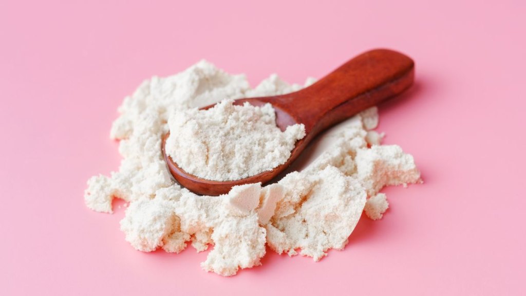 Wooden spoon with protein or collagen powder on pink background close up.