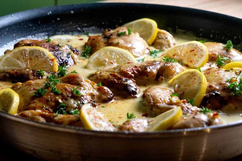 close up of skillet with seared lemon chicken piccata in creamy sauce, with lemon slices