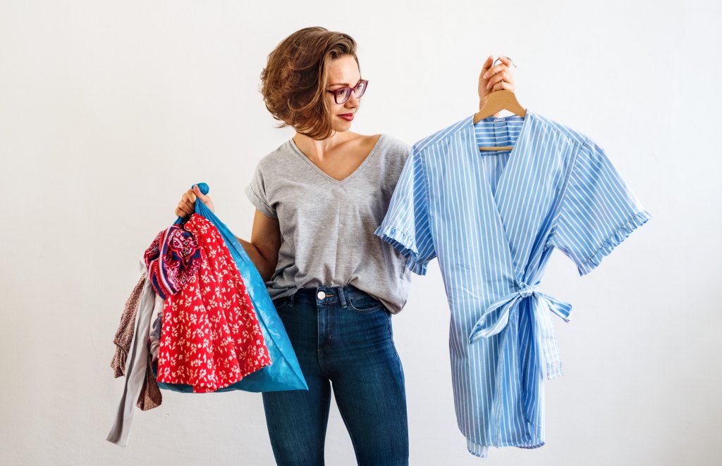 Woman looks at a new piece of clothing on a hanger and holds a bag of old clothes in a donation bag.