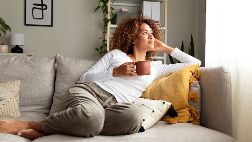 Pensive multiracial woman relaxing at home, sitting on the sofa drinking morning coffee. Lifestyle concept.